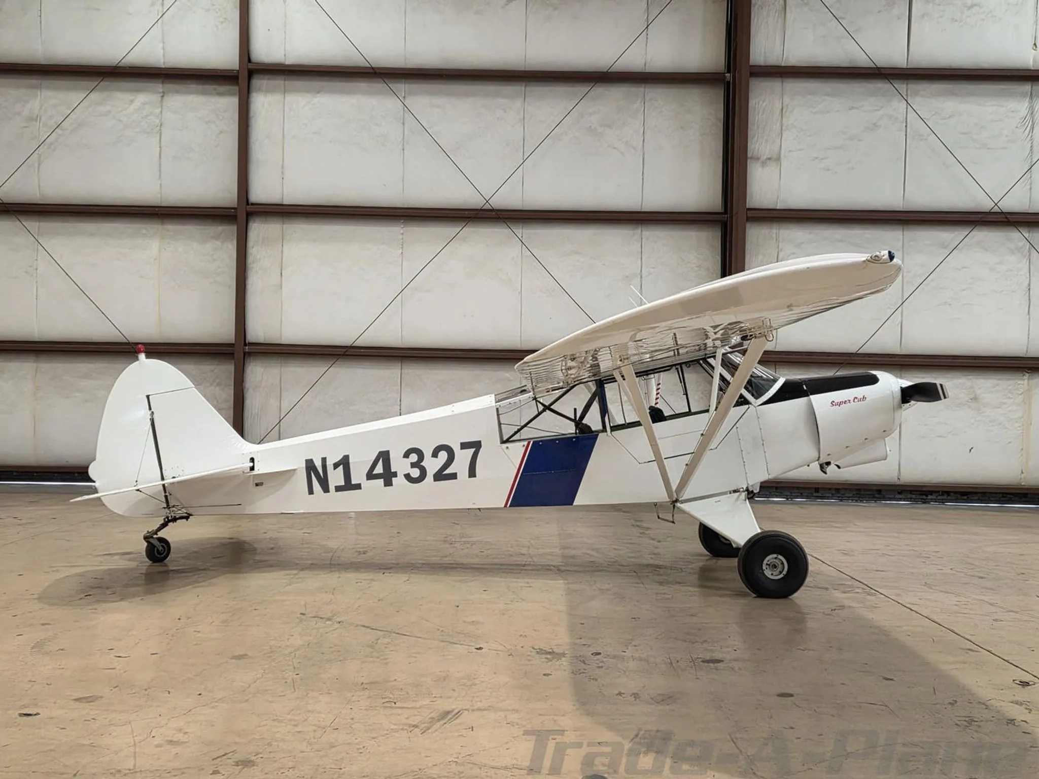 A white Piper Super Cub aircraft with blue and red stripes, registration N14327, viewed from the right side in a hangar.