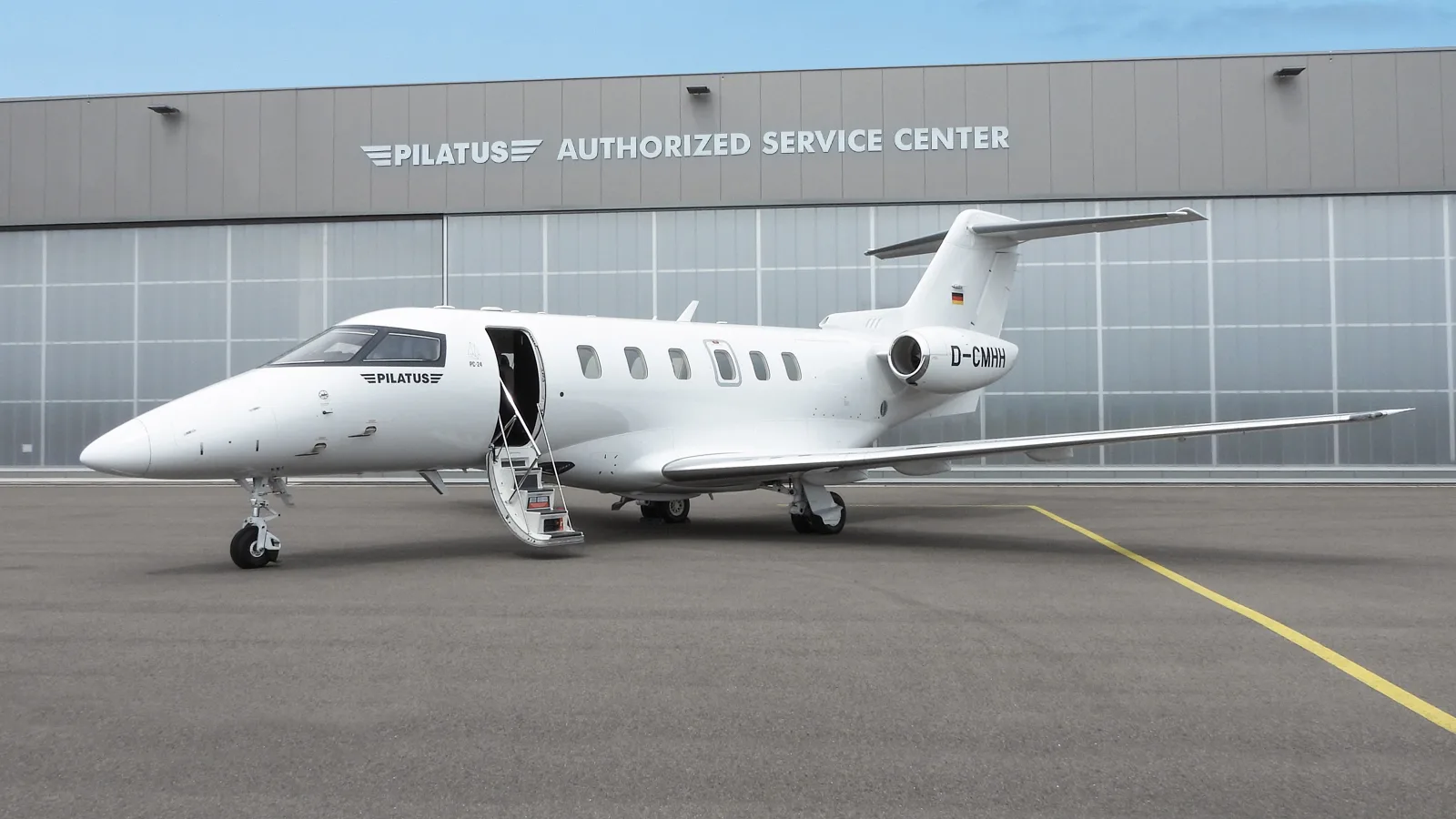 A white Pilatus PC-24 business jet with tail number D-CMHH is parked on an tarmac in front of a Pilatus Authorized Service Center hangar.