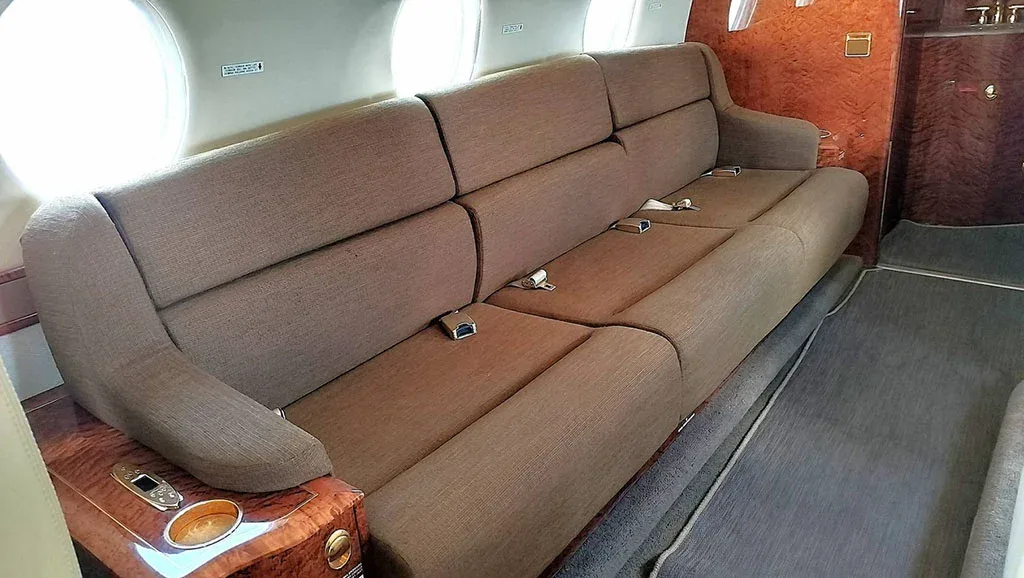 A long, brown fabric sofa-style seat with seatbelts along the side of a private jet cabin, next to a window and wood paneling.