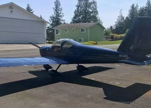 A dark blue single-engine aircraft with tail number N72DJ is parked on a concrete pad with houses and trees in the background.