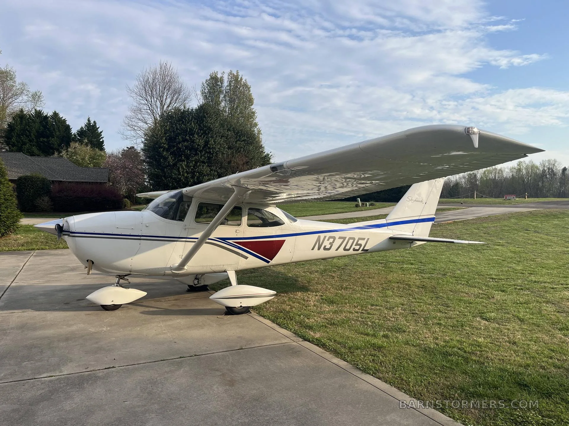 A white Cessna Skyhawk (N3705L) with blue and maroon stripes is parked on a concrete pad next to a grassy area under a partly cloudy sky.