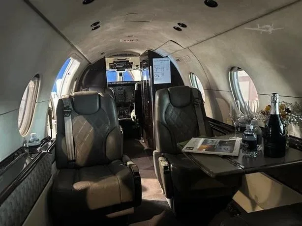 The elegant cabin interior of a business jet, showing two grey leather seats, a table with champagne, and a view towards the cockpit.
