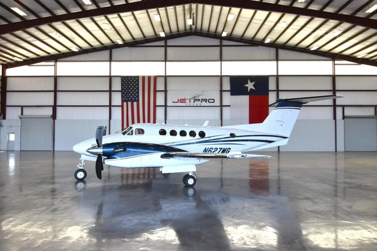 A white and blue Beechcraft King Air N627MG sits in a hangar with American and Texas flags.