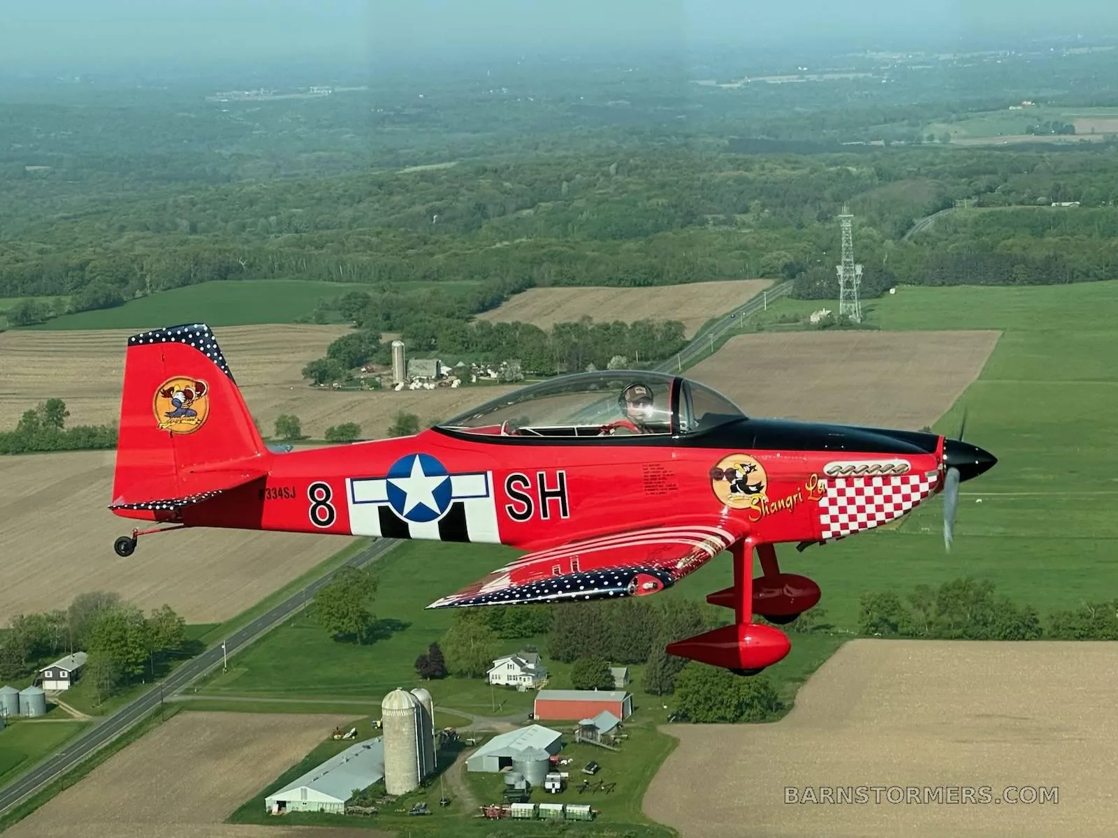 A red single-engine aircraft with a checkered nose and patriotic tail and wing tips flying over green fields.