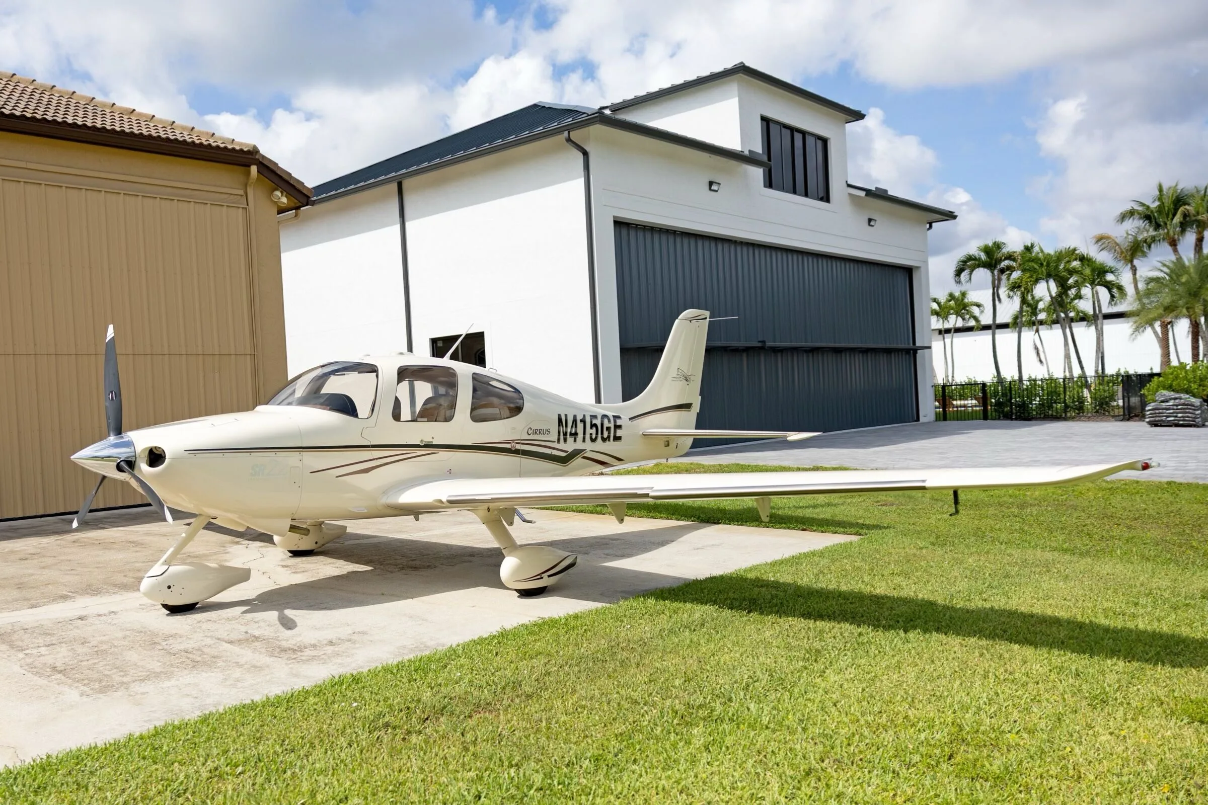 A white Cirrus SR22 aircraft with black and maroon stripes, registration N415GE, parked on a concrete pad next to a white hangar building.