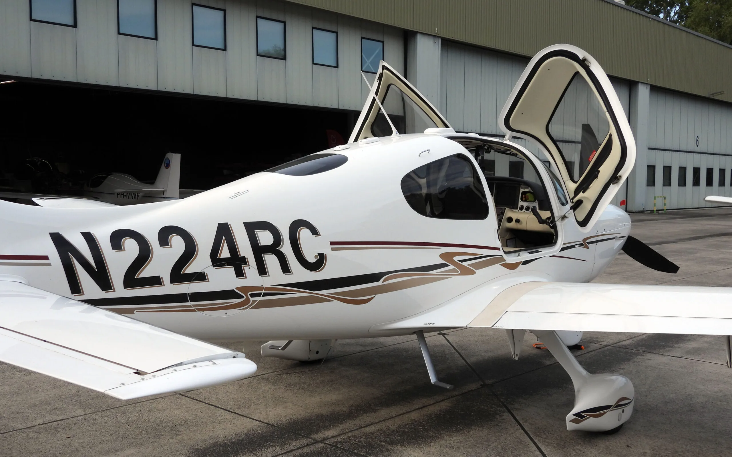 A white Cirrus SR22T (N224RC) with its gull-wing doors open, parked on an apron in front of a hangar.