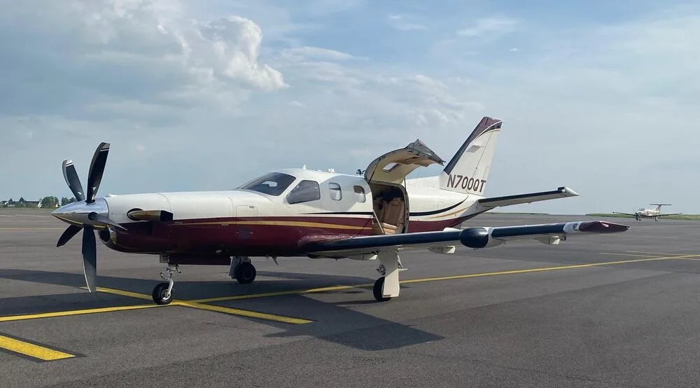 A white and maroon single-engine turboprop aircraft with an open passenger door on an airfield under a cloudy sky.