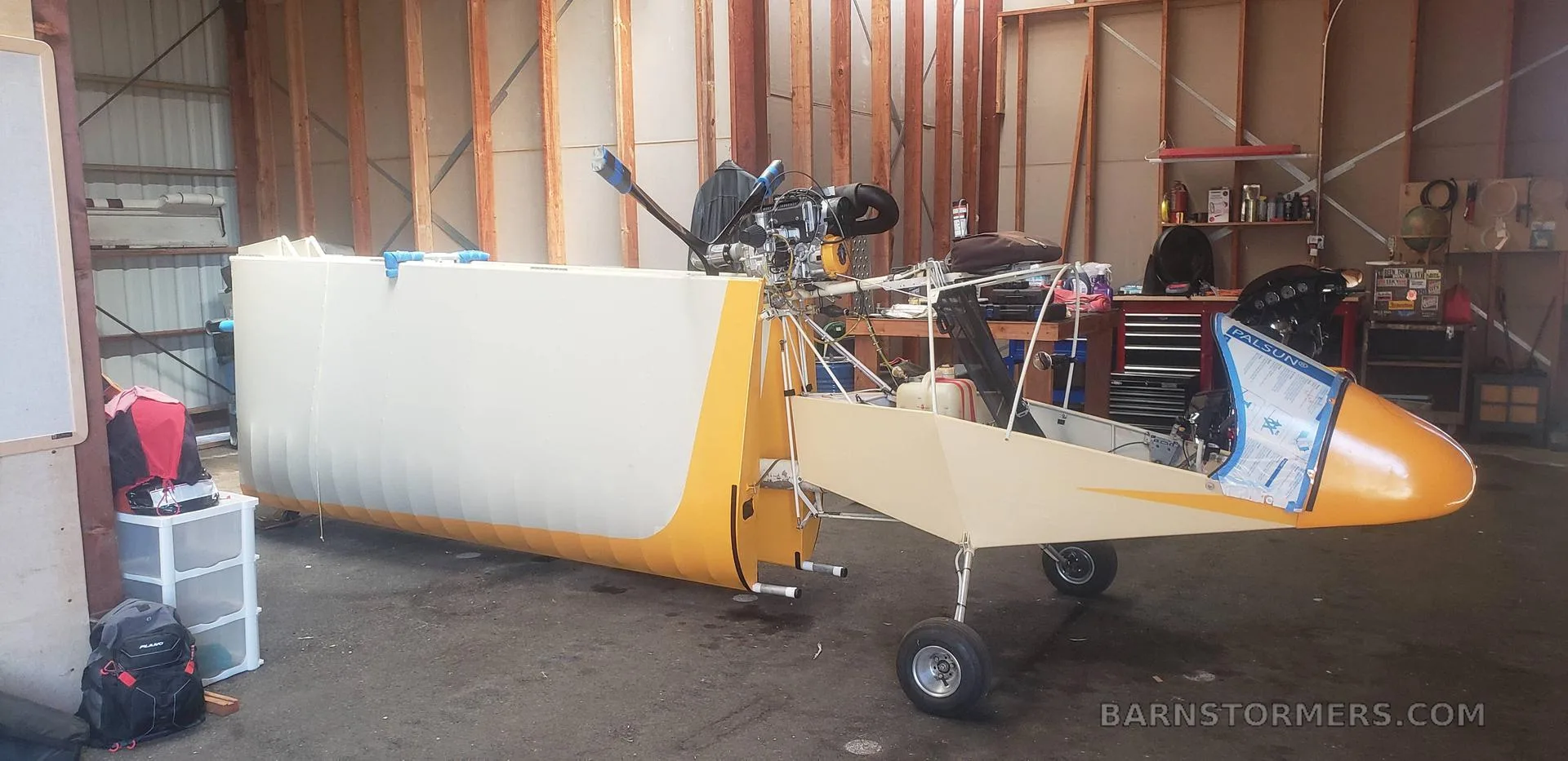 A yellow and white ultralight aircraft, partially assembled with its engine exposed, sits in a workshop.