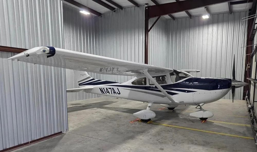 A white and blue Cessna 182 aircraft with registration N147AJ parked inside a metal hangar.