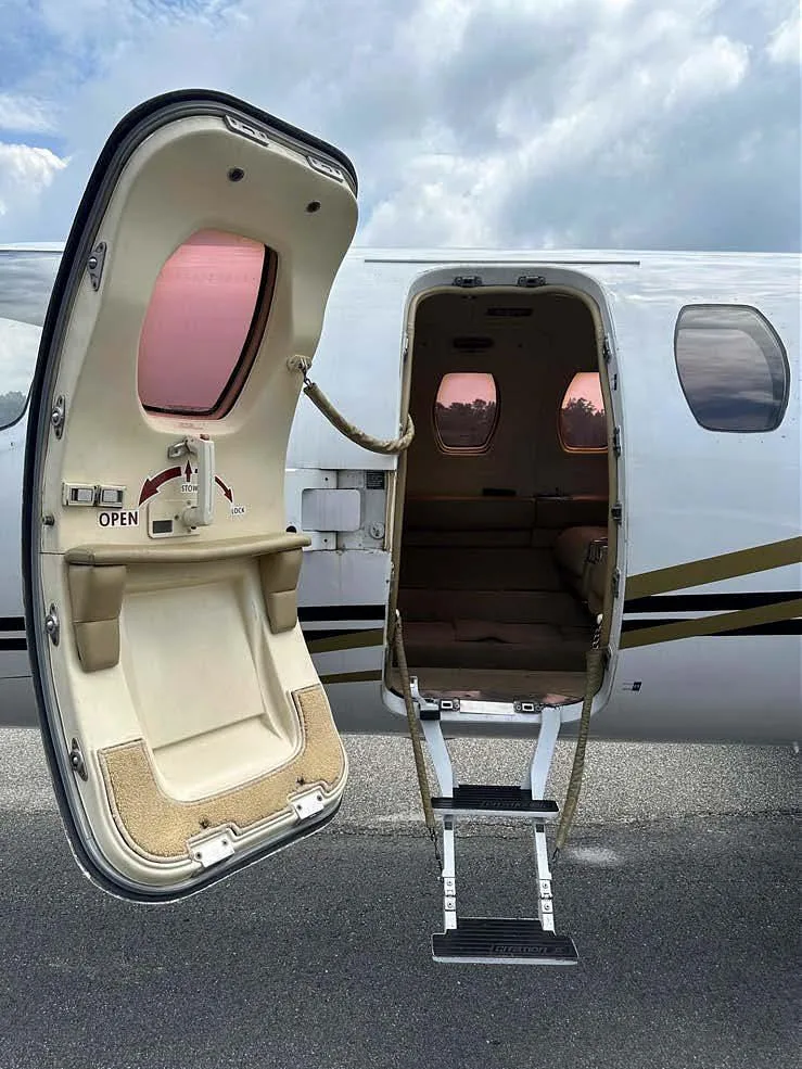 The open entry door and retractable steps of a white business jet, revealing a glimpse of the beige interior.