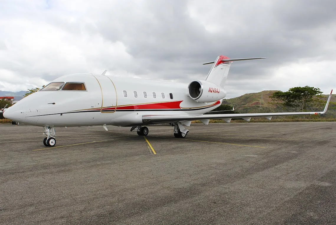 A white Bombardier Challenger business jet with red and gold stripes is parked on an asphalt tarmac under a cloudy sky.