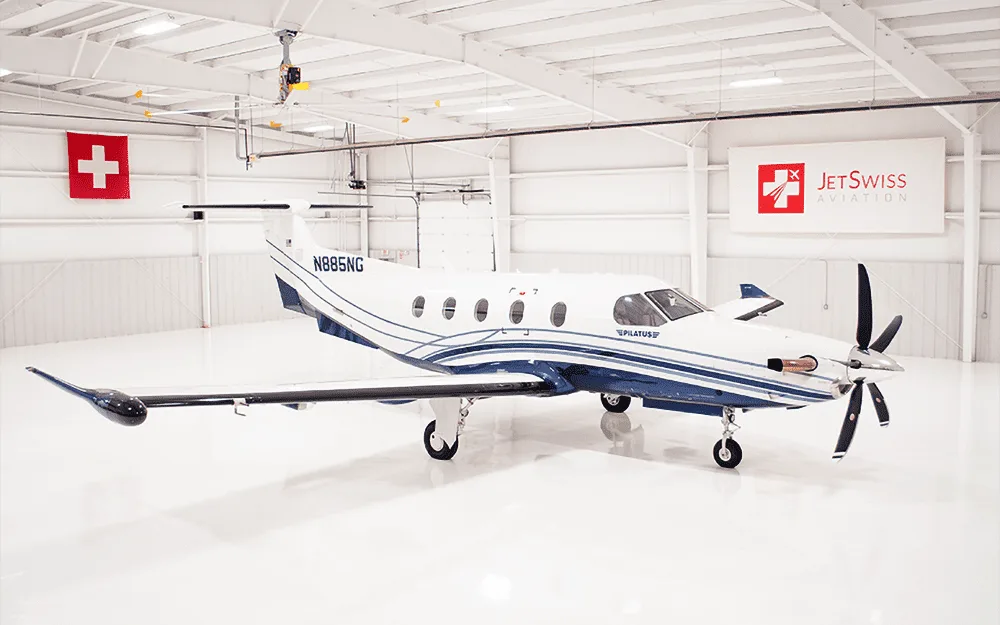 A white and blue Pilatus PC-12 aircraft, tail number N885NG, is parked in a brightly lit hangar, viewed from the front-right.