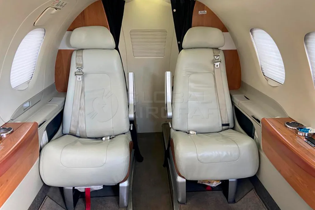 The interior cabin of an Embraer Phenom 100, showing two light grey seats with wooden side tables.