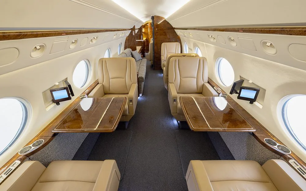 The luxurious cabin interior of a private jet, featuring beige leather seats and polished wood tables.