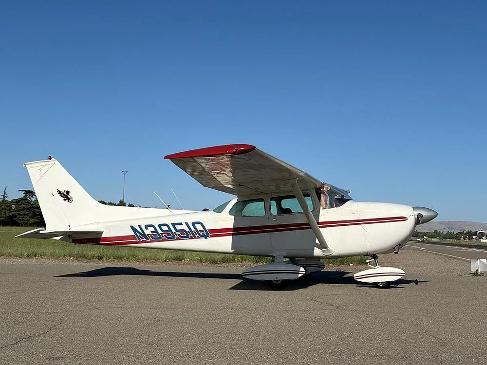 A white Cessna aircraft with red stripes and N3951Q registration parked on an airfield with a clear blue sky and distant trees.