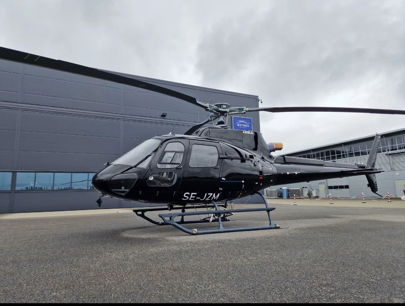 A black Airbus helicopter, registration SE-JZM, parked on an asphalt apron in front of a dark grey hangar under an overcast sky.