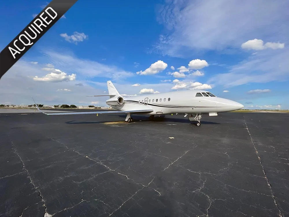 A white business jet with a dark stripe is parked on an asphalt tarmac under a blue sky with scattered clouds.