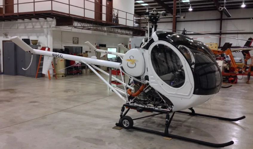 A white helicopter with a black canopy and exposed engine components is parked in a hangar.