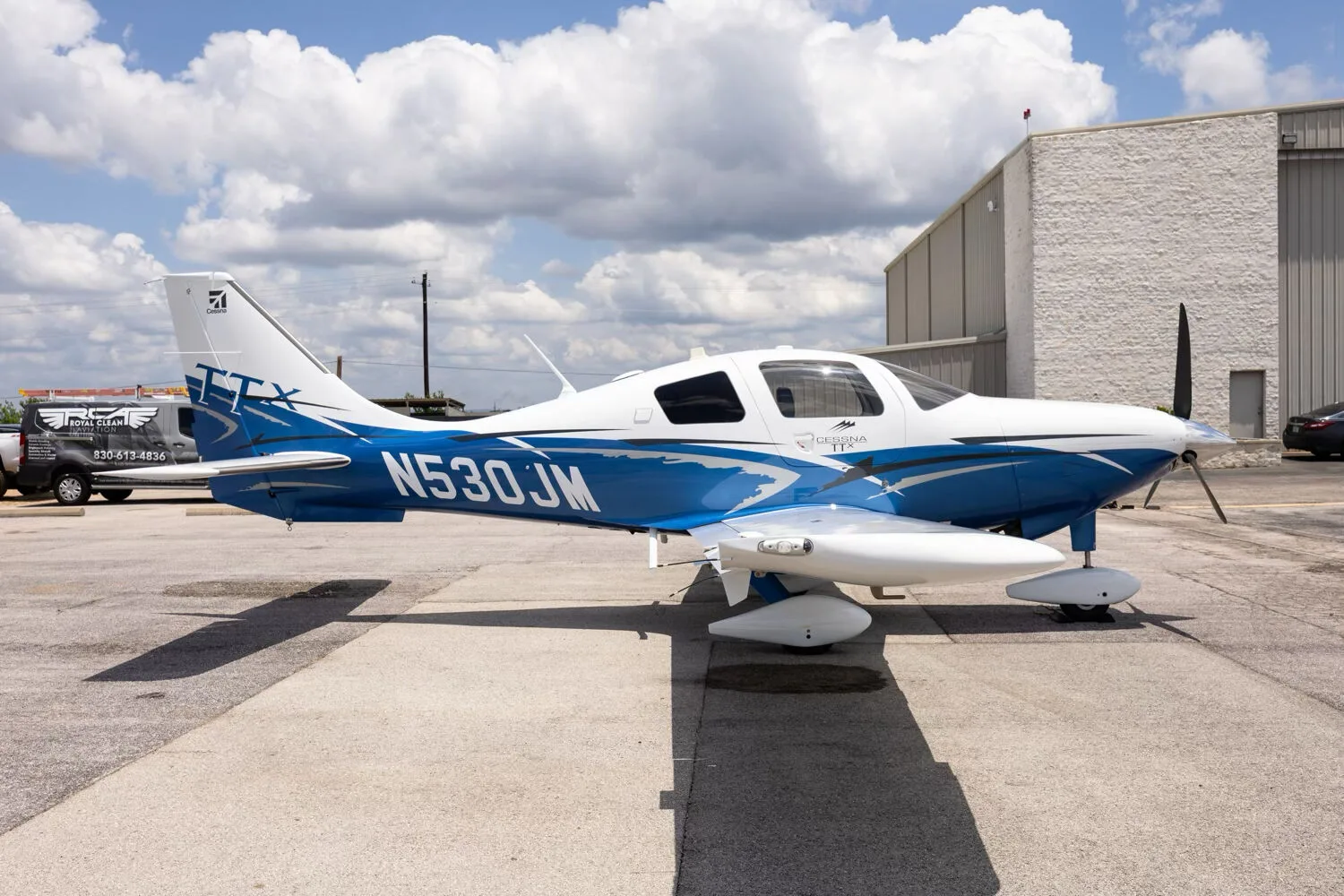 A side view of the white and blue Cessna TTX N530JM parked on a tarmac next to a hangar.