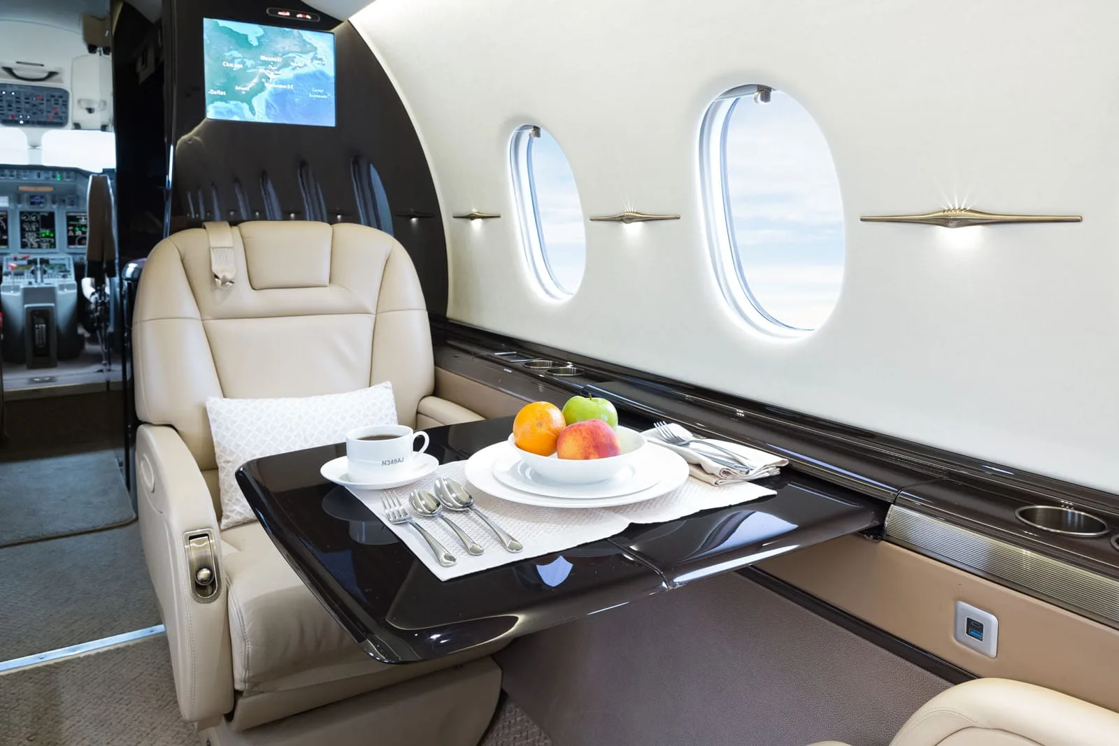 A luxurious aircraft interior with a beige leather seat, a set table with fruit and coffee, and a map displayed on a screen in the background.
