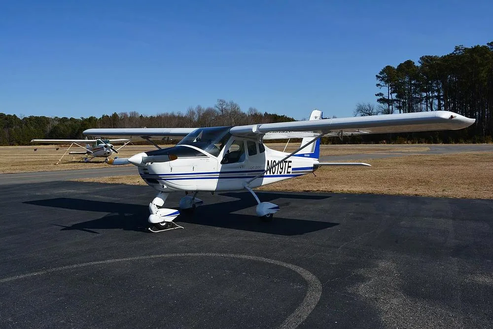 A white Tecnam Light Sport aircraft with blue and grey stripes parked on an asphalt ramp with a grassy field in the background.