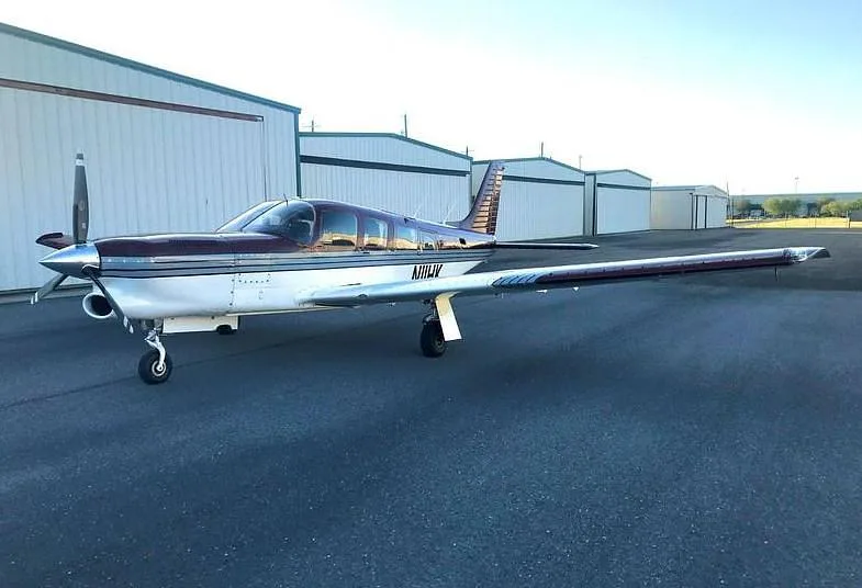 A full side view of the burgundy and white single-engine aircraft, N11HK, parked on an airfield next to hangars.