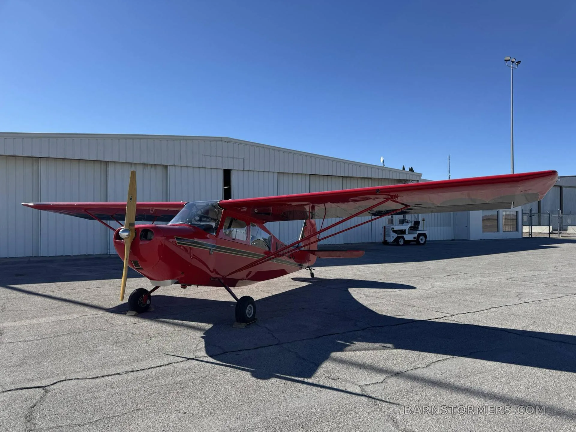 A red high-wing monoplane with a wooden propeller is parked on an asphalt ramp next to a white hangar under a clear blue sky.