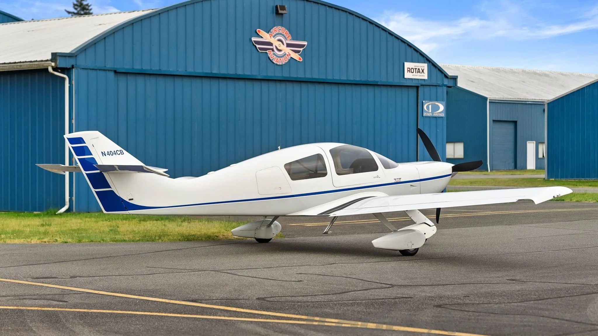 A side view of a white single-engine aircraft with blue stripes on the fuselage and tail, registration N404CB, parked on an asphalt runway in front of a blue hangar.