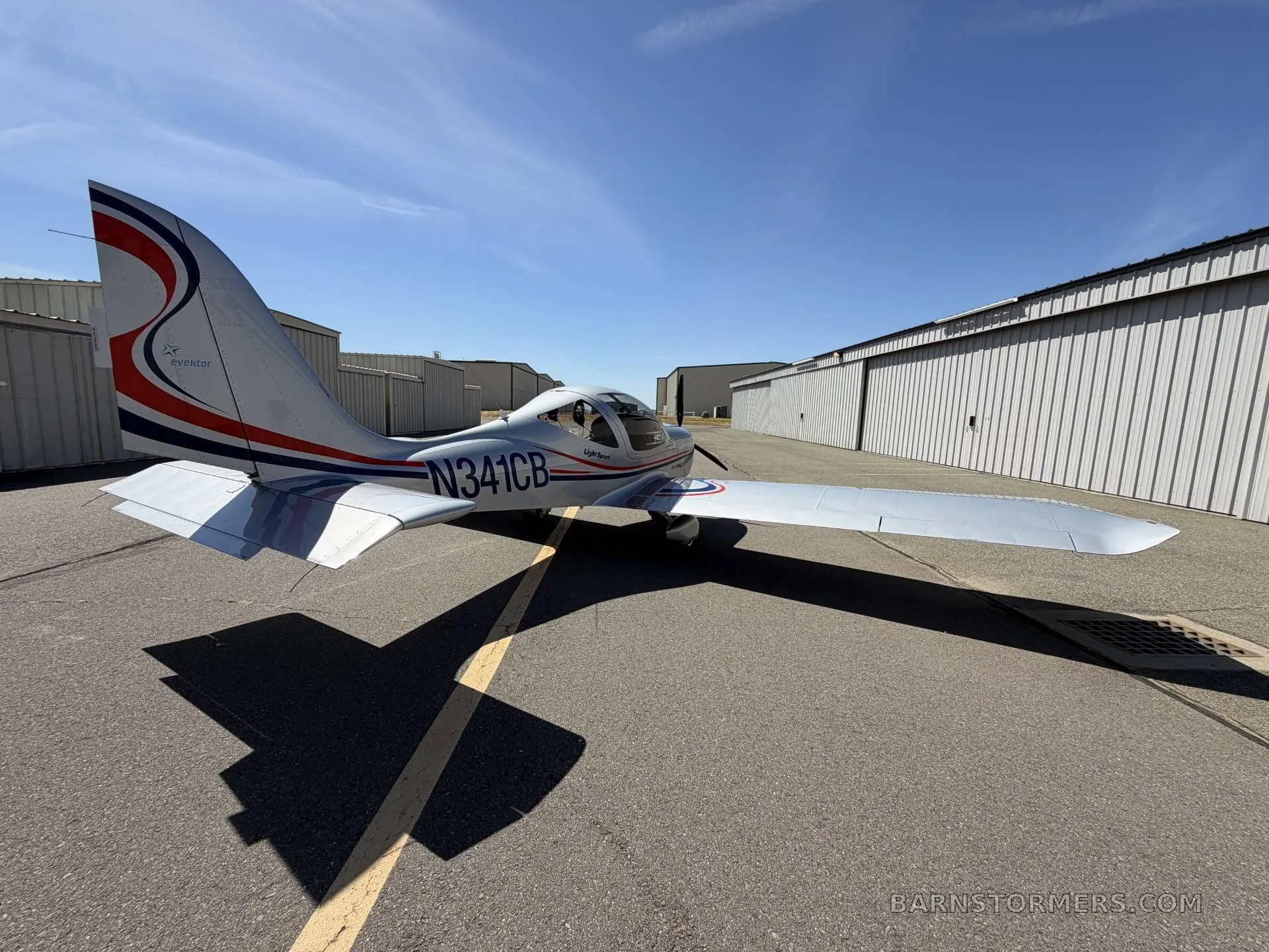 A rear-right view of the silver SportStar MAX Turbo aircraft with tail number N341CB and red and blue stripes, parked on an asphalt tarmac with hangars in the background.