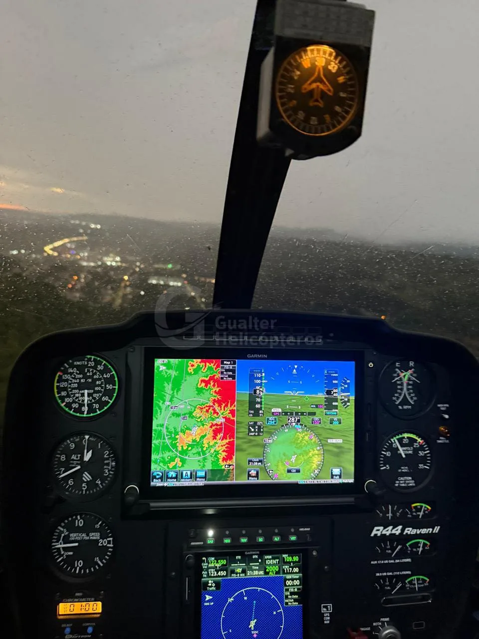 The cockpit of a Robinson R44 Raven II helicopter at night, showing the instrument panel and city lights visible through the windshield.