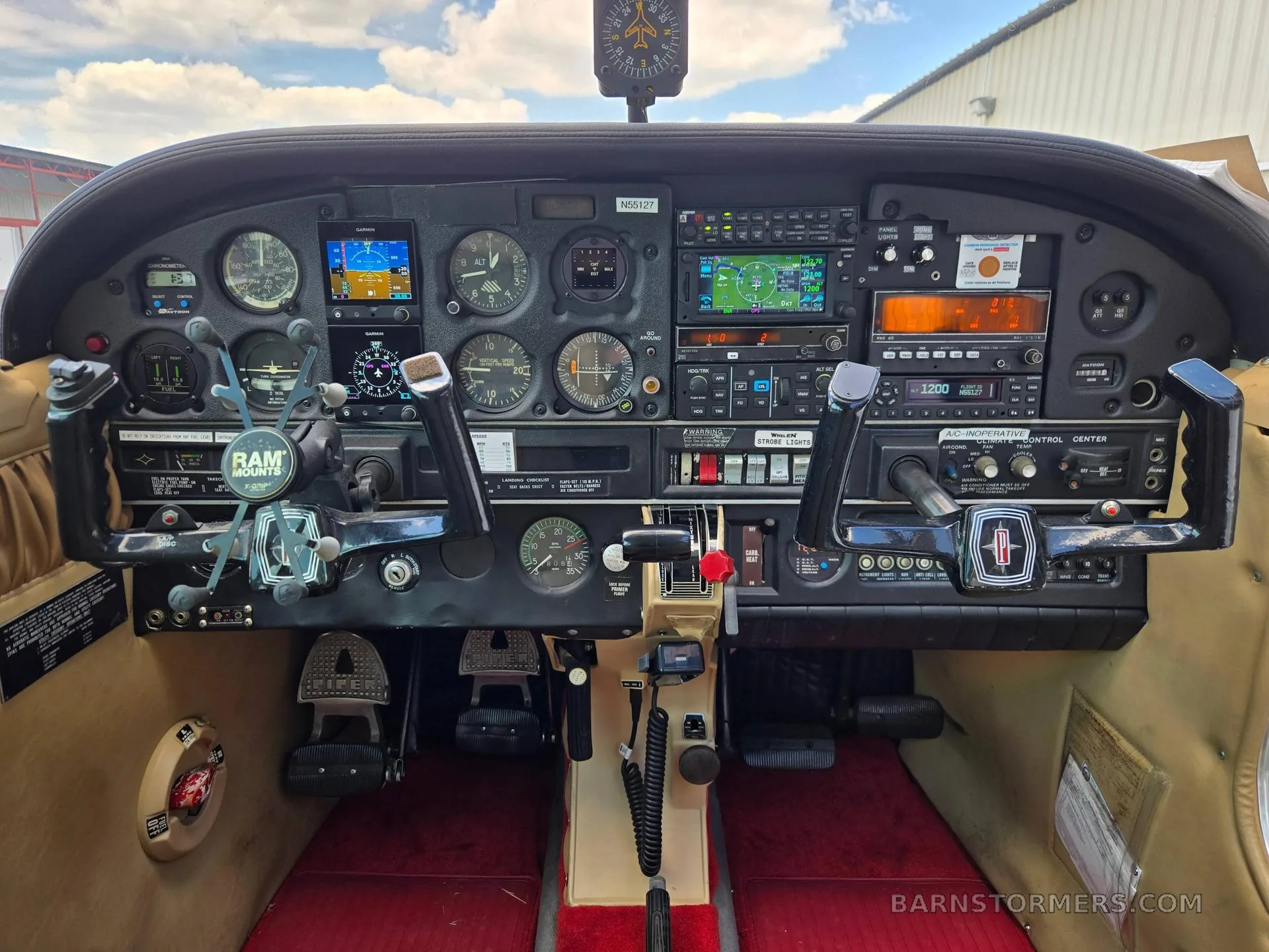 The detailed instrument panel of a small aircraft features multiple gauges, two Garmin displays, and dual yokes with a red carpeted floor below.