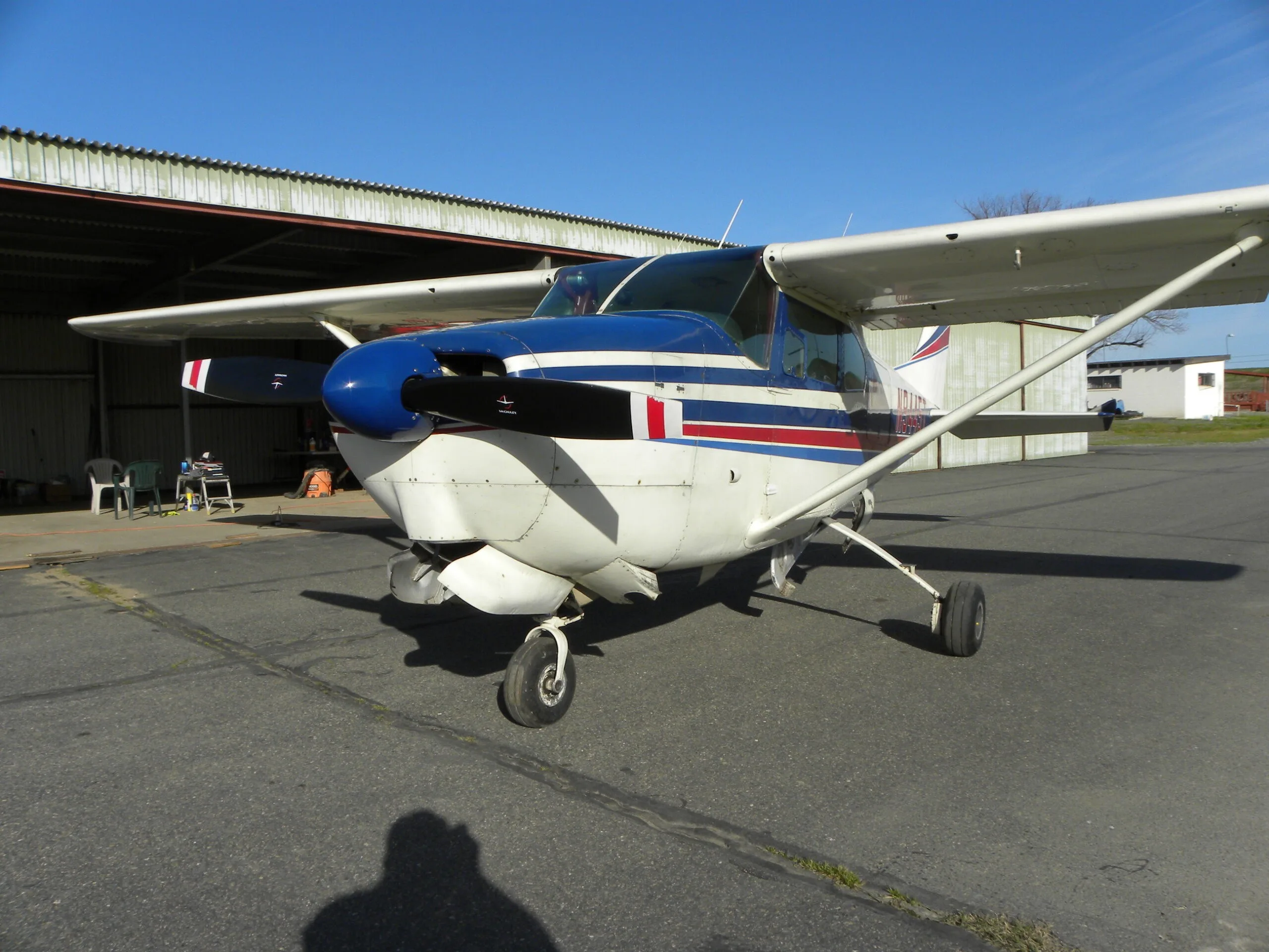A white Cessna 182 with blue and red stripes parked on a tarmac in front of a hangar.