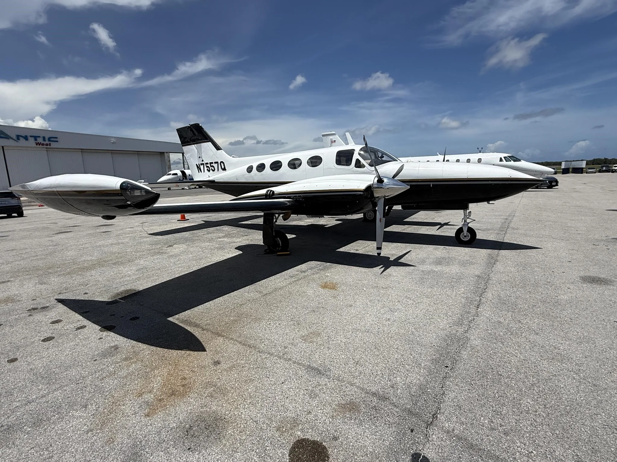 A side view of the white and black twin-engine propeller aircraft N7557Q on an airfield under a partly cloudy sky.