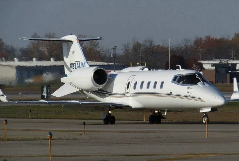 A white business jet with blue and gold stripes, registration N824LM, parked on a tarmac with buildings in the background.