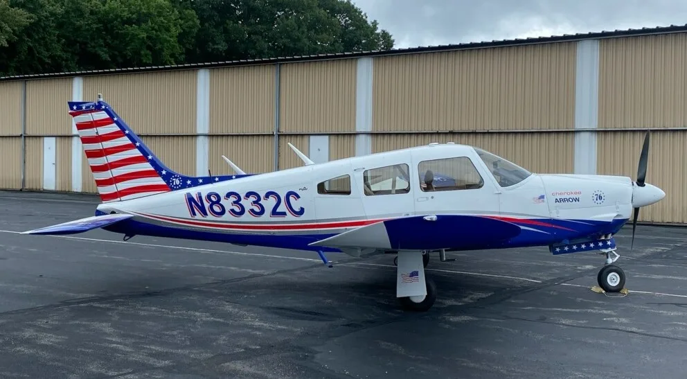 A white and blue Piper Cherokee Arrow with an American flag tail sits on an asphalt ramp in front of a hangar.