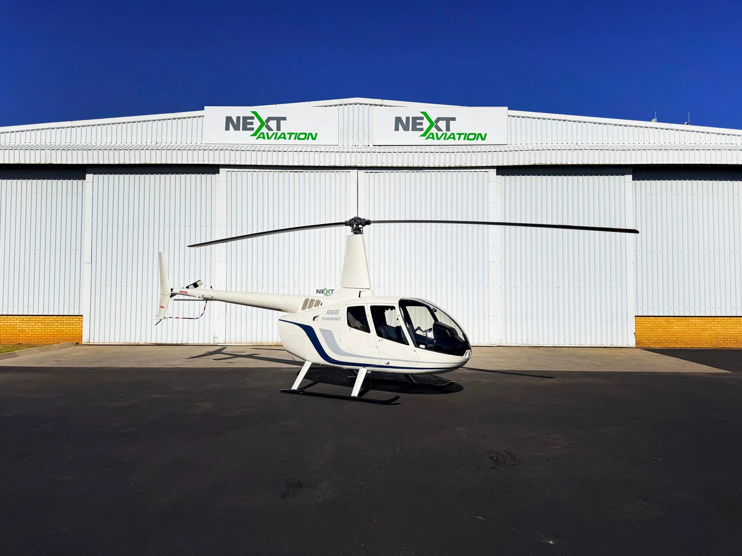 A white Robinson R66 Turbine helicopter with blue stripes parked on a tarmac in front of a hangar.