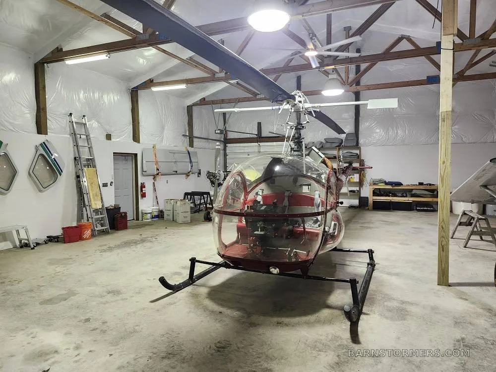 A red and white helicopter with a clear bubble canopy is parked in a hangar with a small plane in the background.