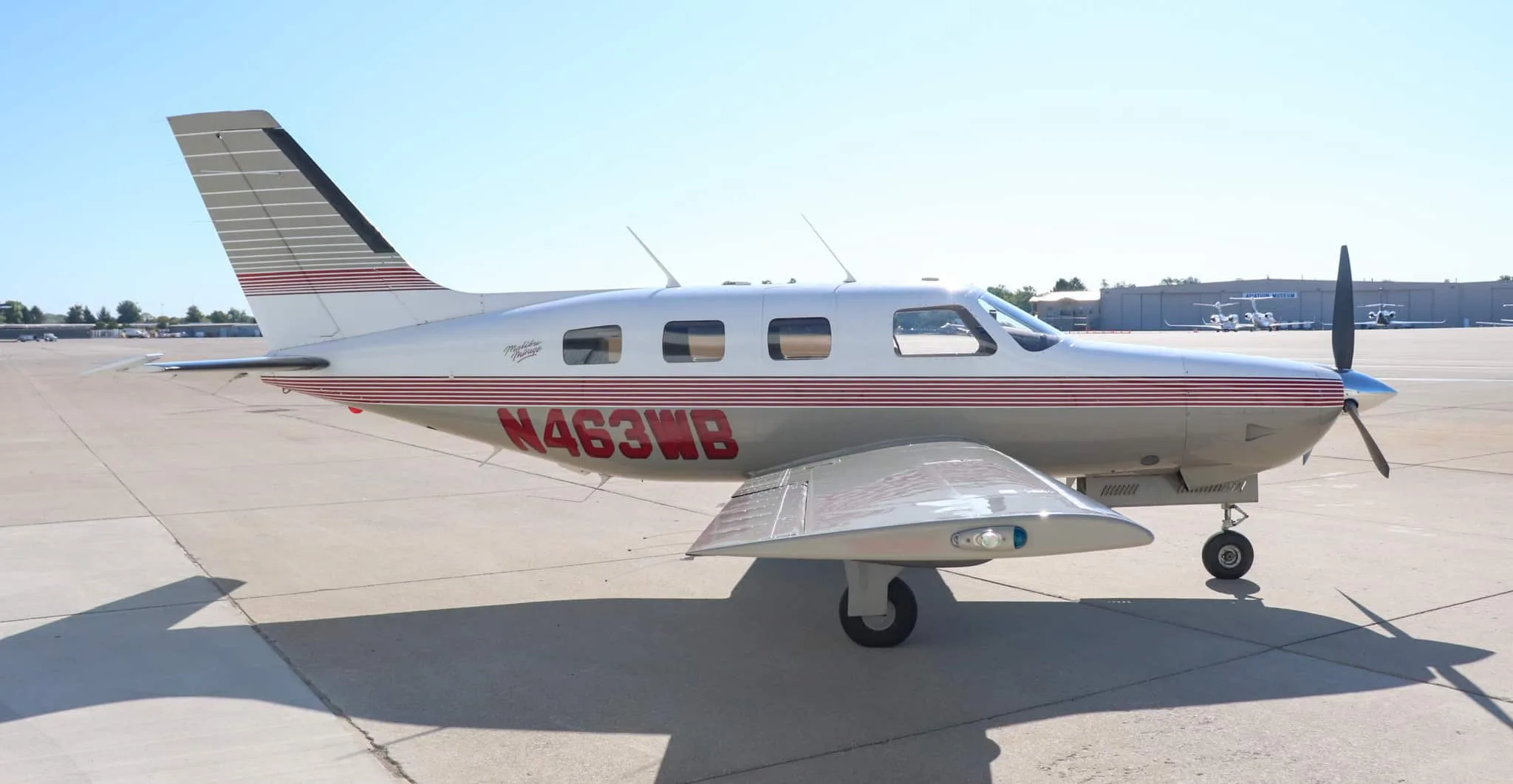 A side view of the Piper Malibu Mirage N463WB, showing its white and tan paint scheme with red stripes, parked on an airport tarmac.