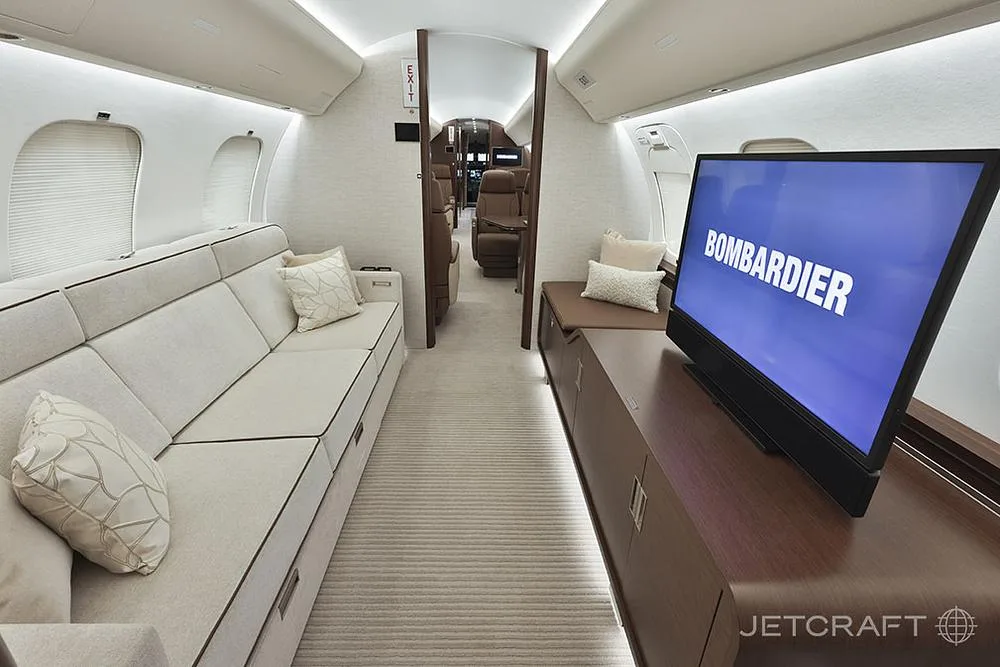 A business jet cabin interior features a light-colored sofa, a large screen displaying 'BOMBARDIER', and a view towards the rear.