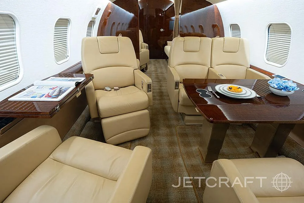 The private jet cabin interior with beige seats, a wooden table set with a plate and flowers, and magazines on a side table.