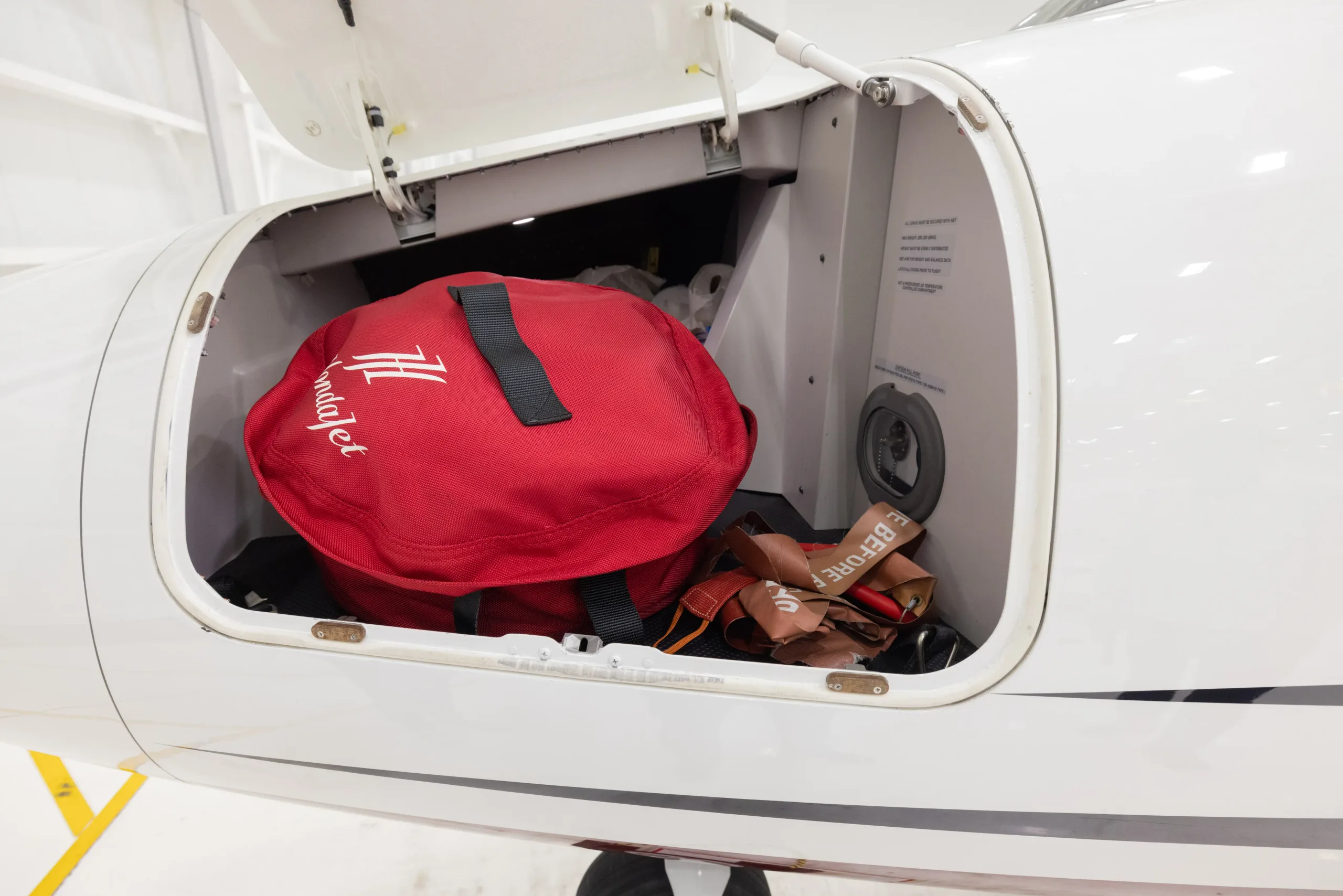 The open forward baggage compartment of a HondaJet, revealing a red bag and a 'REMOVE BEFORE FLIGHT' tag.