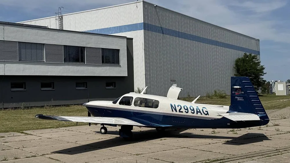 A Mooney Ovation 2 Platinum Edition aircraft, N299AG, with a blue and white paint scheme, parked on concrete next to a building.