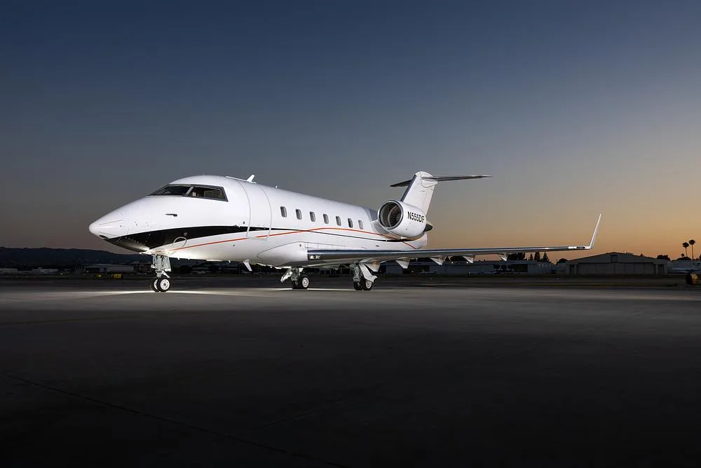 A white Bombardier Challenger 605 business jet, registration N555DF, is parked on an airport tarmac at dusk.