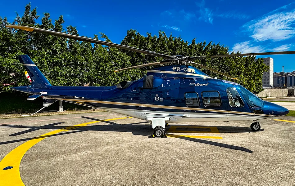 A dark blue and white Agusta Grand helicopter with a gold stripe is parked on a concrete helipad under a clear blue sky.