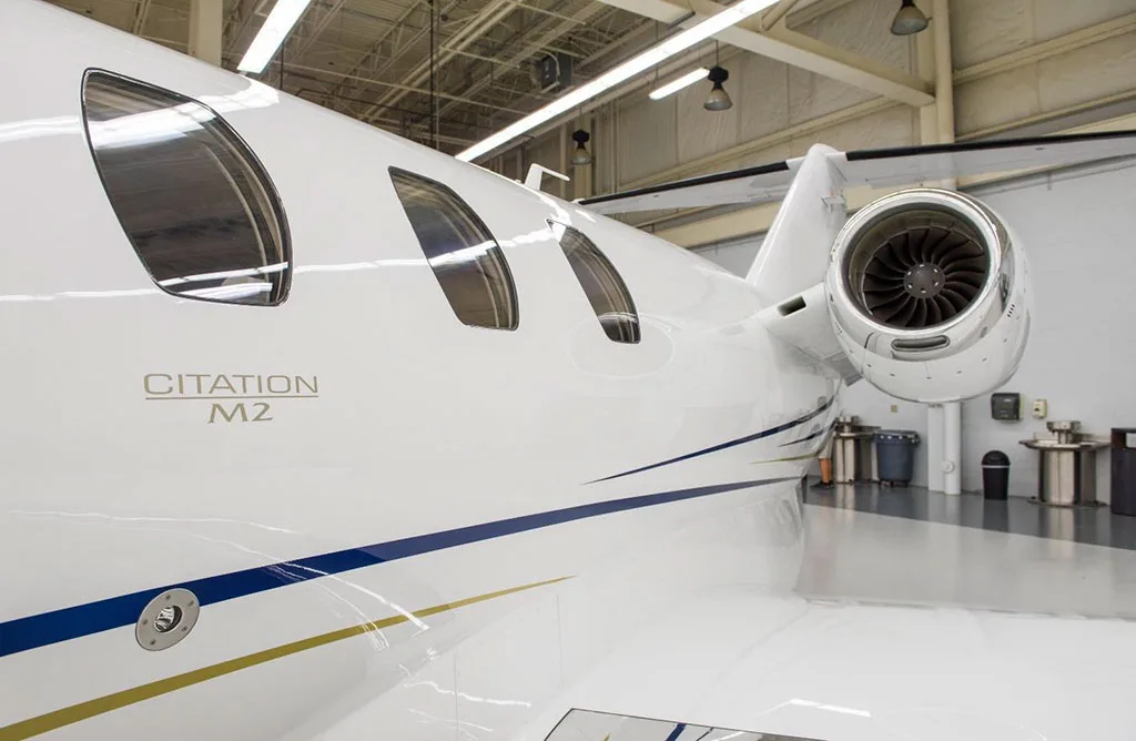 A close-up view of the rear fuselage and engine of a white Citation M2 jet with blue and gold stripes, inside a hangar.