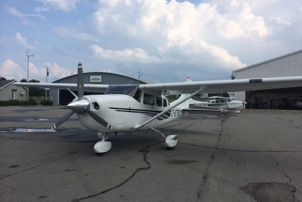 A white single-engine propeller aircraft, registration C-GTMQ, parked on an asphalt tarmac with other aircraft and hangars in the background.