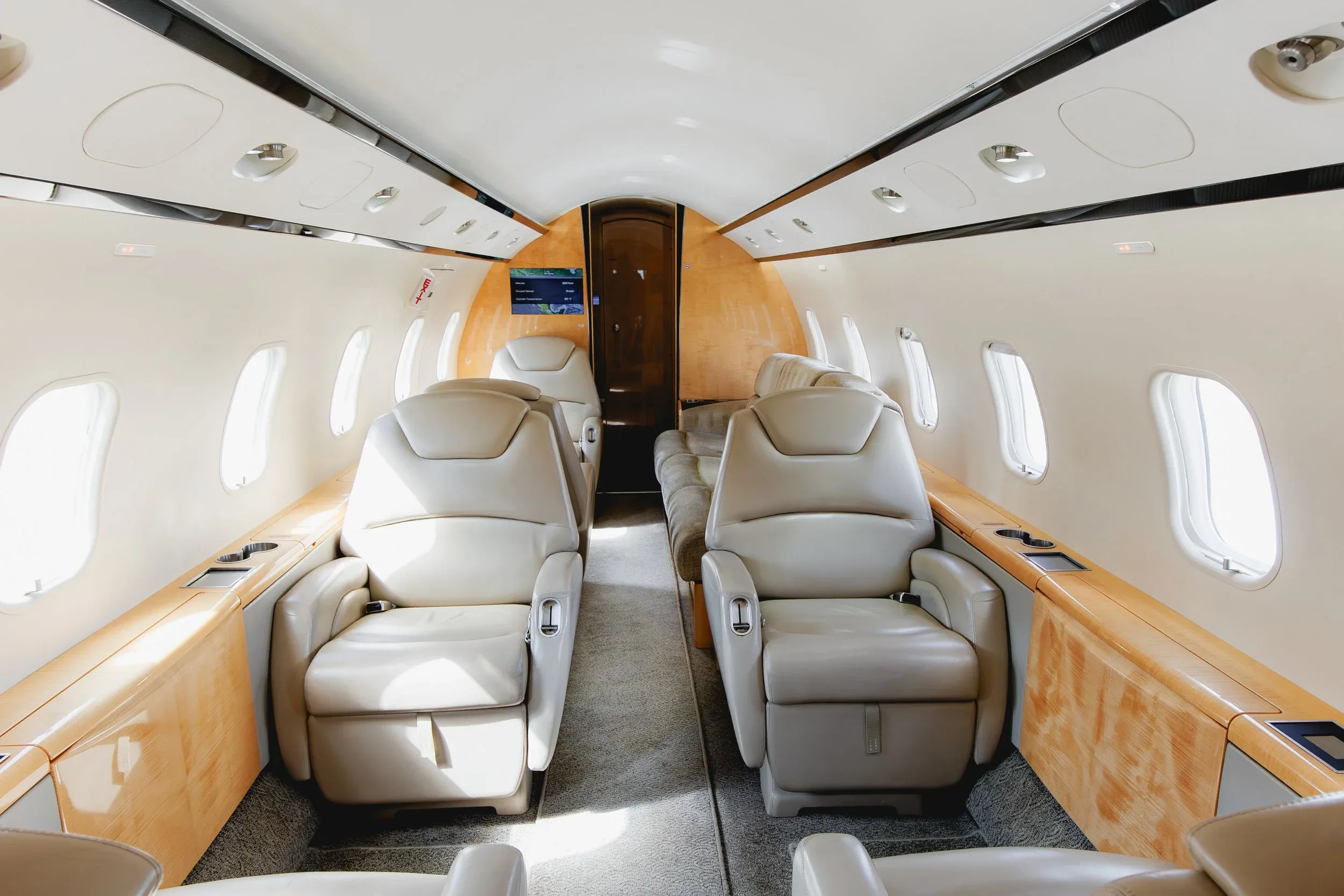 The interior of a private jet cabin with light beige leather seats and light wood paneling, looking towards the rear.
