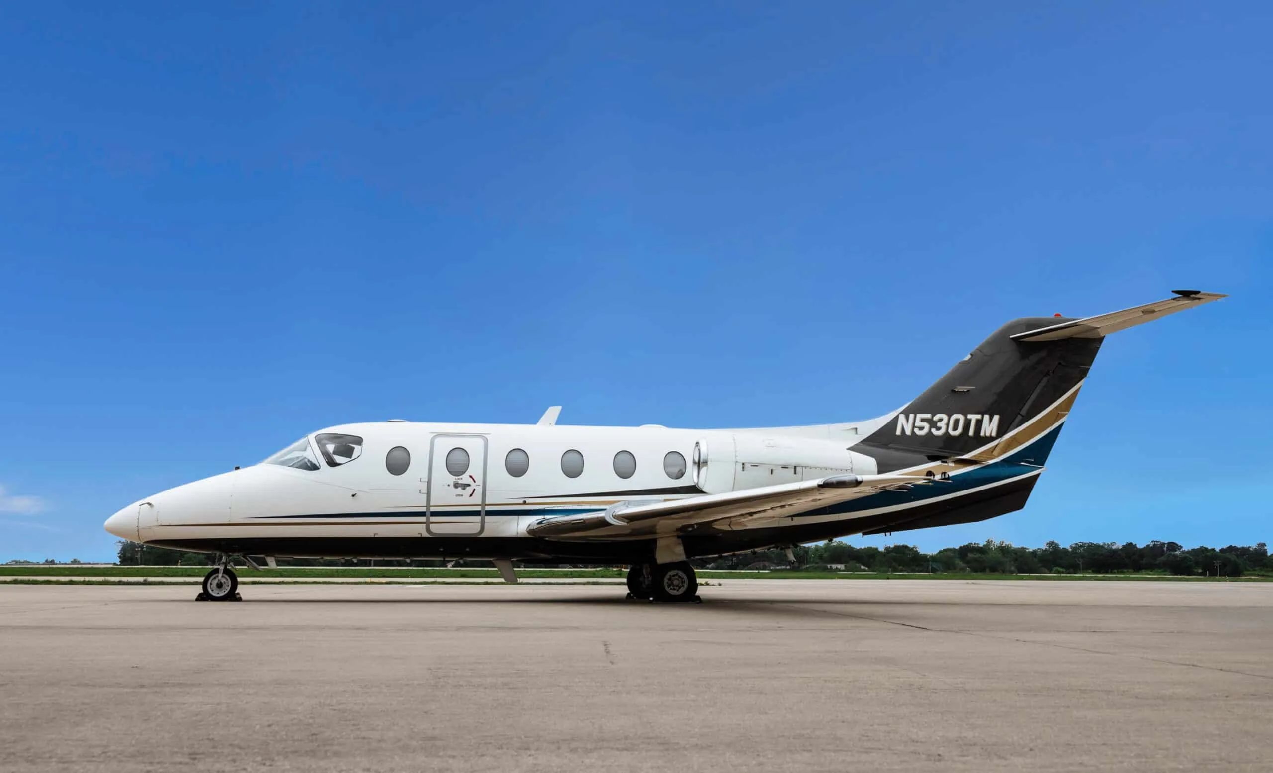 A white, black, and gold private jet (N530TM) is parked on an airfield under a clear blue sky.