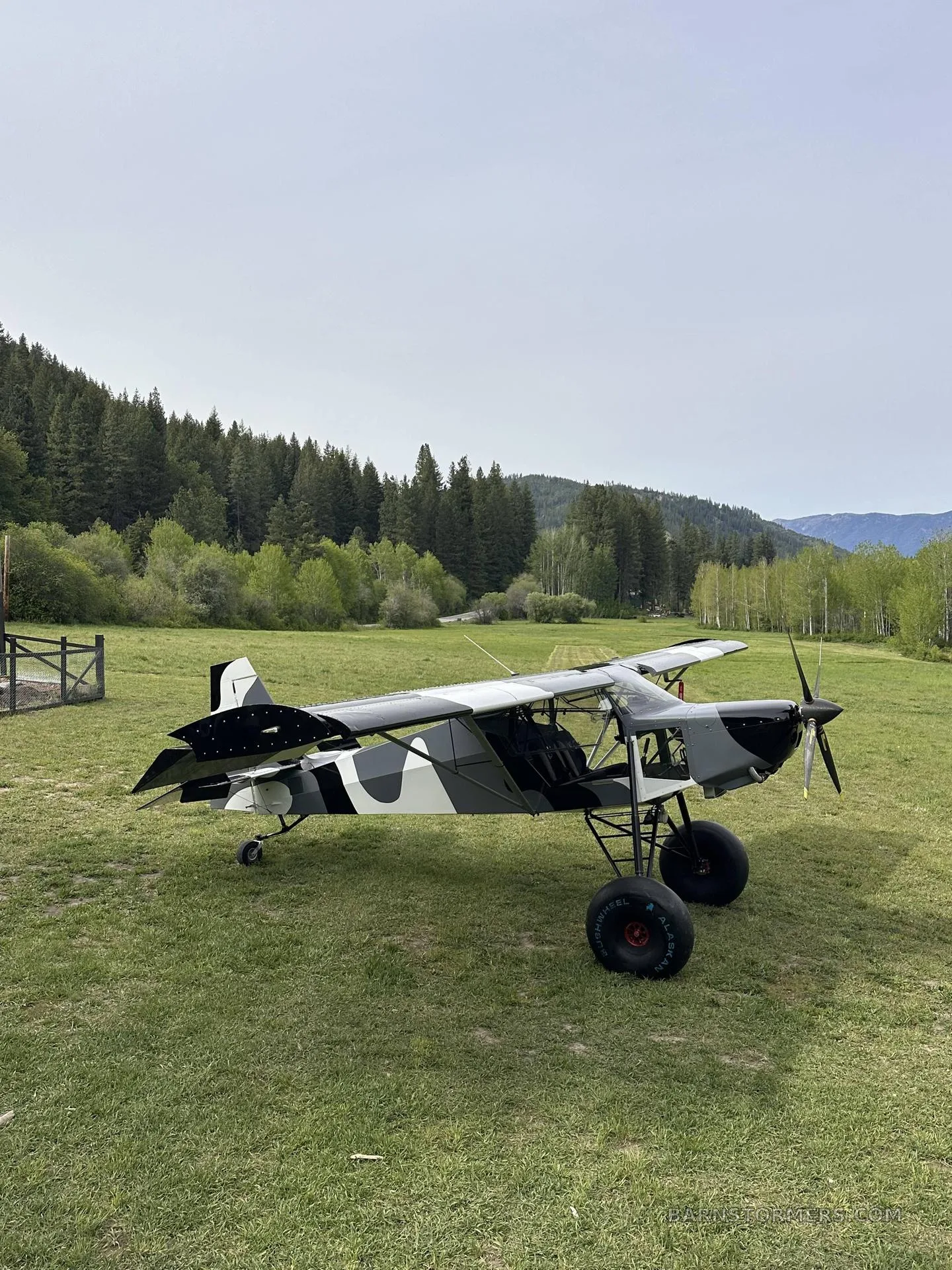 A black, white, and gray camouflage STOL aircraft with large tundra tires is parked on a grassy field, facing a distant runway strip.