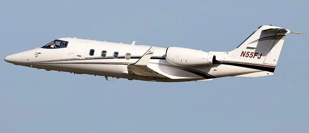 A white Learjet 60 with brown and black stripes, tail number N55FJ, flying against a clear blue sky.